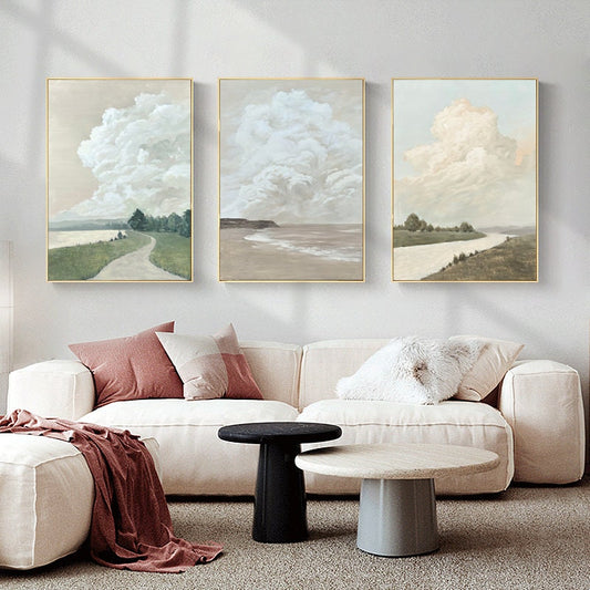 May include: Three framed canvas prints with a minimalist landscape theme. Each print features a large, fluffy cloud in a pale blue, white, or cream color against a light beige background. The bottom of each print shows a different landscape scene: a path through a grassy field, a beach with waves, and a river winding through a field.
