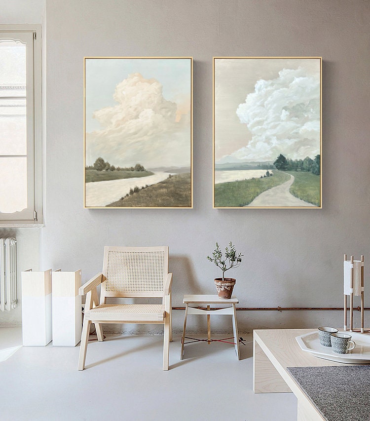 May include: Two framed landscape paintings with a muted color palette. The paintings depict a river winding through a grassy field with a large fluffy cloud in the sky. The paintings are hung on a light gray wall.