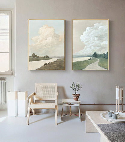 May include: Two framed landscape paintings with a muted color palette. The paintings depict a river winding through a grassy field with a large fluffy cloud in the sky. The paintings are hung on a light gray wall.