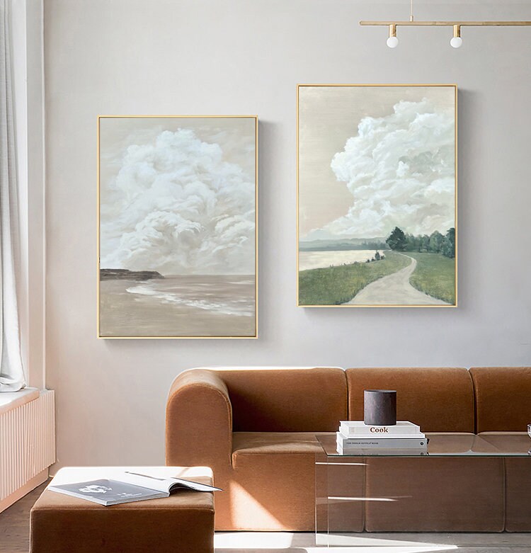 May include: Two abstract landscape paintings with a minimalist style. The paintings feature a cloudy sky and a path leading to a body of water. The paintings are framed in gold and hung on a white wall.