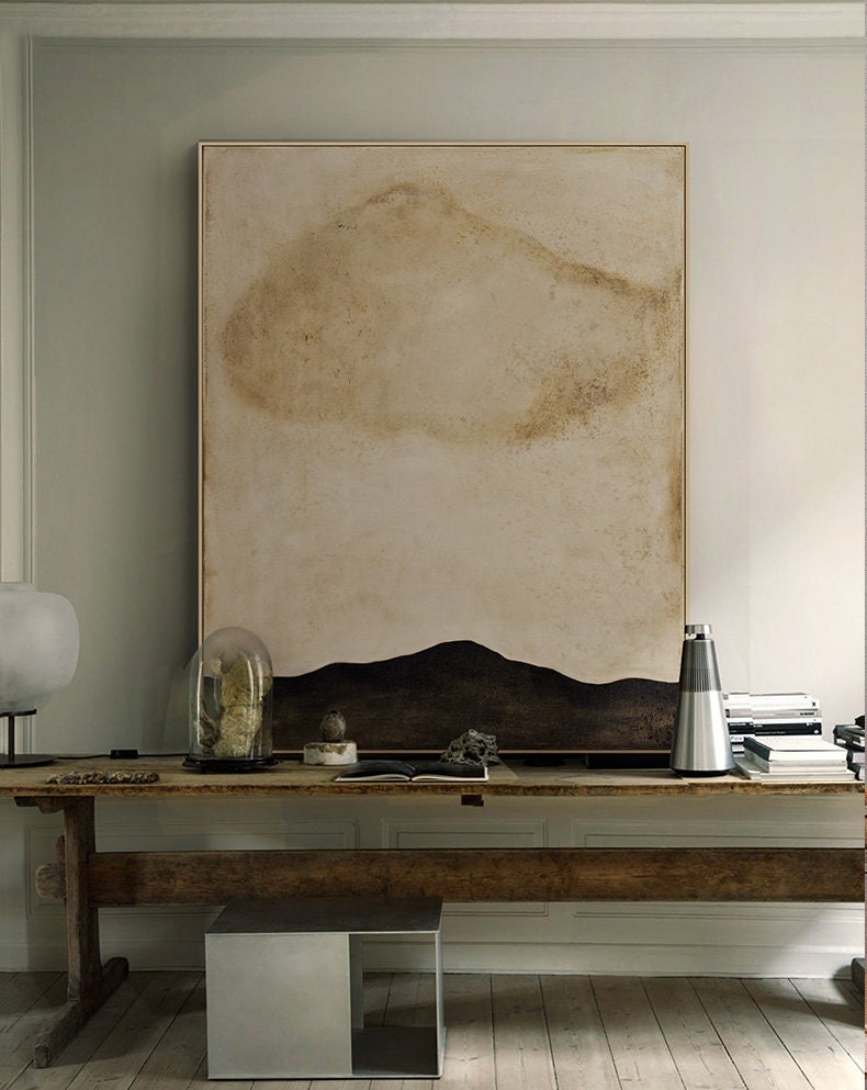 May include: A large abstract painting with a brown and beige background and a black mountain range at the bottom. The painting is hanging on a white wall above a wooden table with a silver speaker, a glass dome, and a book.