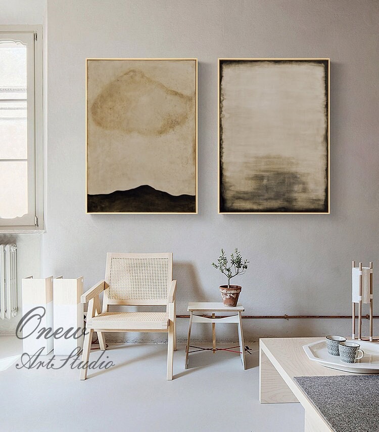 May include: Two abstract paintings in gold frames, one with a brown cloud shape and a black mountain, the other with a brown and gray gradient. The paintings are hanging on a light gray wall above a white wicker chair and a small wooden table with a potted plant.