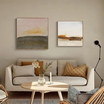 May include: Two abstract landscape paintings in muted tones of brown, gray, and white hang above a beige sofa with brown and beige throw pillows. A vase with white flowers sits on a wooden coffee table in front of the sofa.