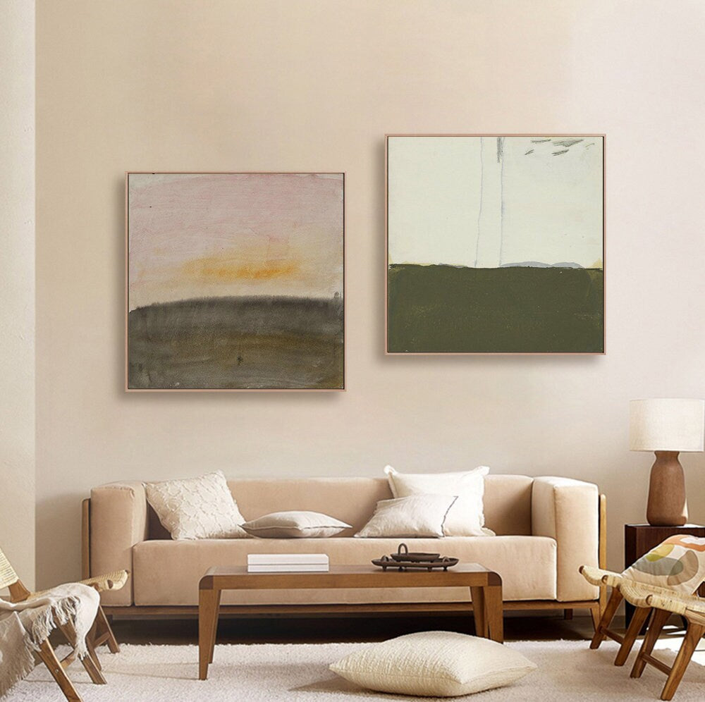 May include: Two abstract landscape paintings in muted tones of pink, green, and brown. The paintings are framed and hung on a beige wall above a beige sofa.
