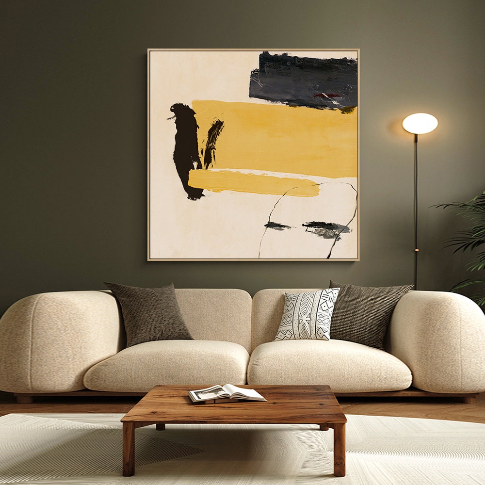 May include: Abstract art print with yellow, black, and white paint strokes on a beige background. The print is framed in a thin, light wood frame and is hanging on a dark green wall above a beige sofa.