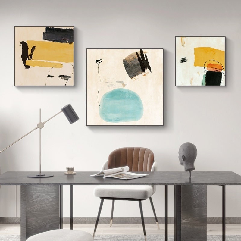 May include: Three abstract paintings with a minimalist style. The paintings feature geometric shapes and brushstrokes in shades of yellow, blue, black, and white. The paintings are hung on a white wall above a desk with a chair.