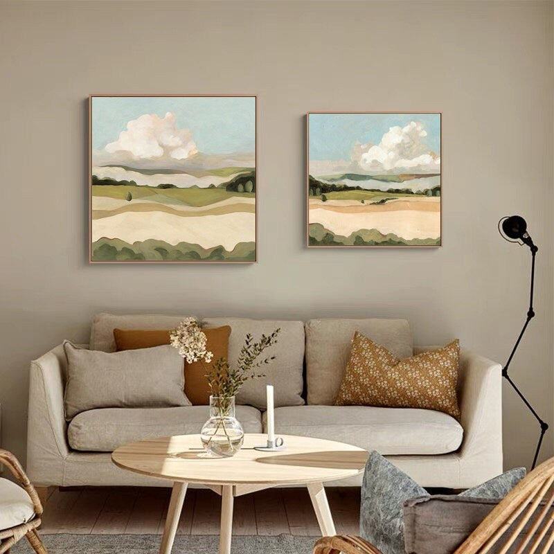 May include: Two abstract landscape paintings with a light blue sky, white clouds, and green hills. The paintings are framed in a light wood frame and are hanging on a light beige wall above a light beige sofa with brown throw pillows.