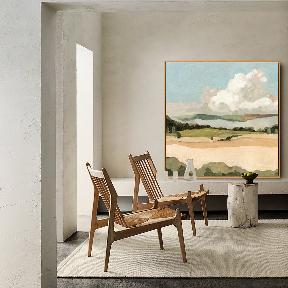May include: A large abstract landscape painting with a white cloud in a blue sky, green hills, and a brown field. The painting is hanging on a wall in a room with two wooden chairs and a wooden table.