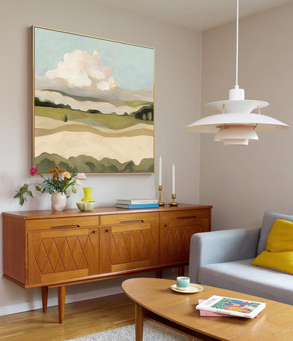 May include: A mid-century modern living room with a light wood sideboard, a gray sofa with a yellow throw pillow, and a coffee table with books and a cup. A painting of a landscape with a large white cloud hangs on the wall above the sideboard. A white pendant light hangs from the ceiling.