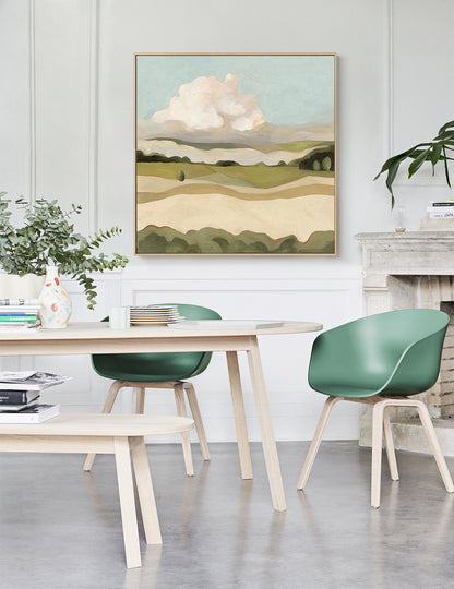 May include: A painting of a landscape with rolling hills, a large white cloud, and a blue sky. The painting is framed in a light wood frame and is hanging on a white wall.
