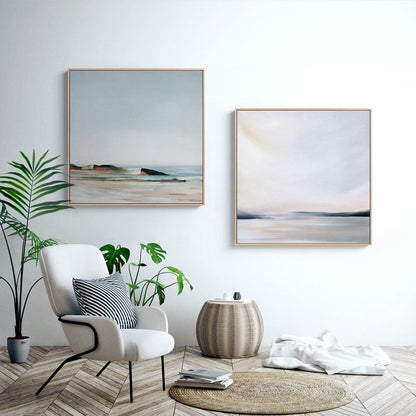 May include: Two abstract landscape paintings in muted tones of blue, gray, and white, hanging on a white wall above a white armchair with a black and white striped pillow. A potted plant is on the left side of the image, and a woven basket-style side table is on the right.