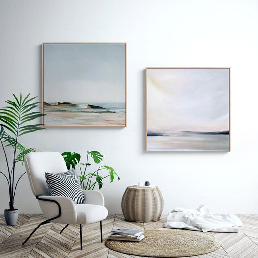 May include: Two abstract landscape paintings in muted tones of blue, gray, and white, hanging on a white wall above a white armchair with a black and white striped pillow. A potted plant is on the left side of the image, and a woven basket-style side table is on the right.