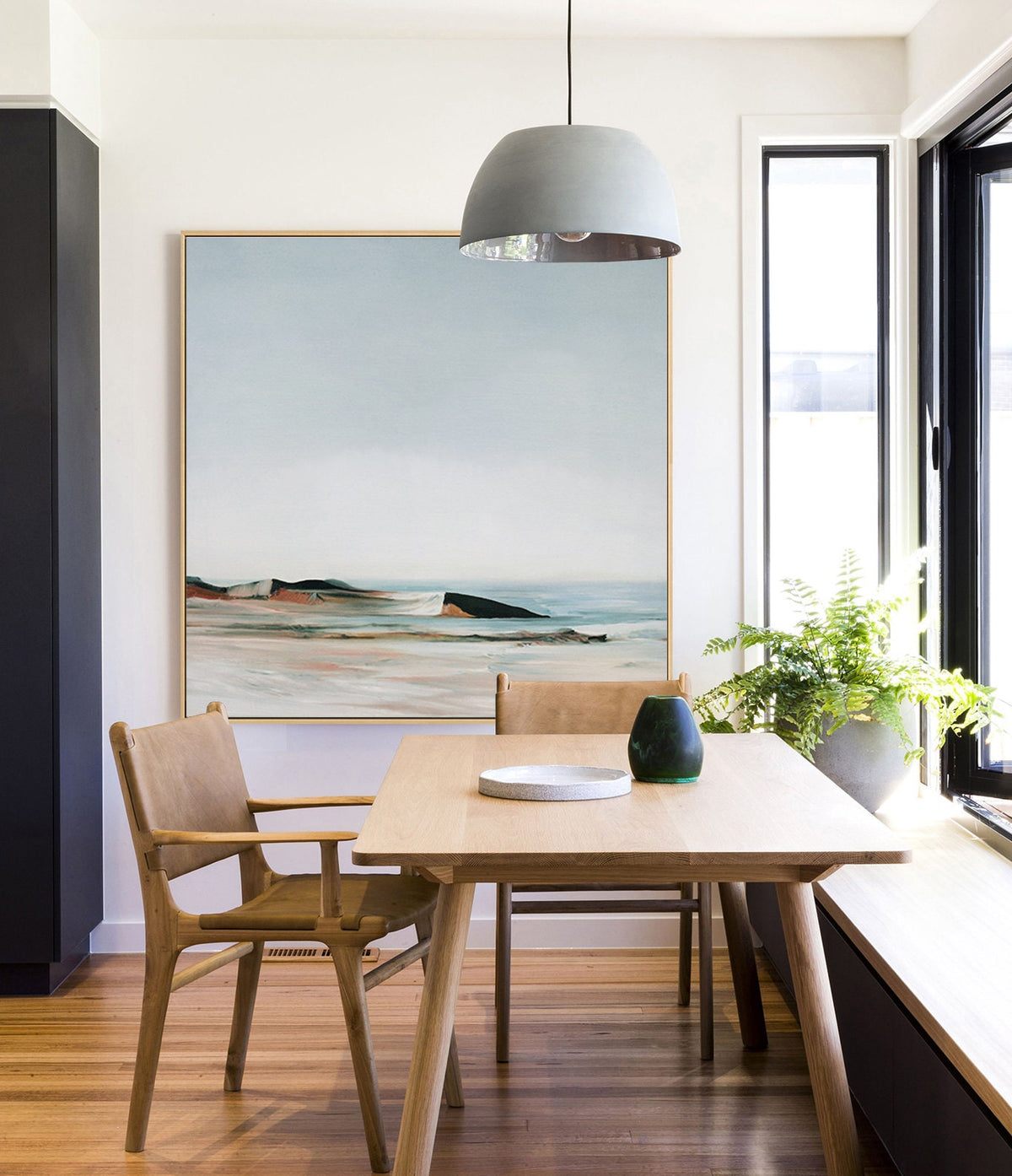 May include: A light wood dining table with two chairs and a gray pendant light hanging above. The table has a white plate and a green vase on it. A painting of a seascape hangs on the wall behind the table.