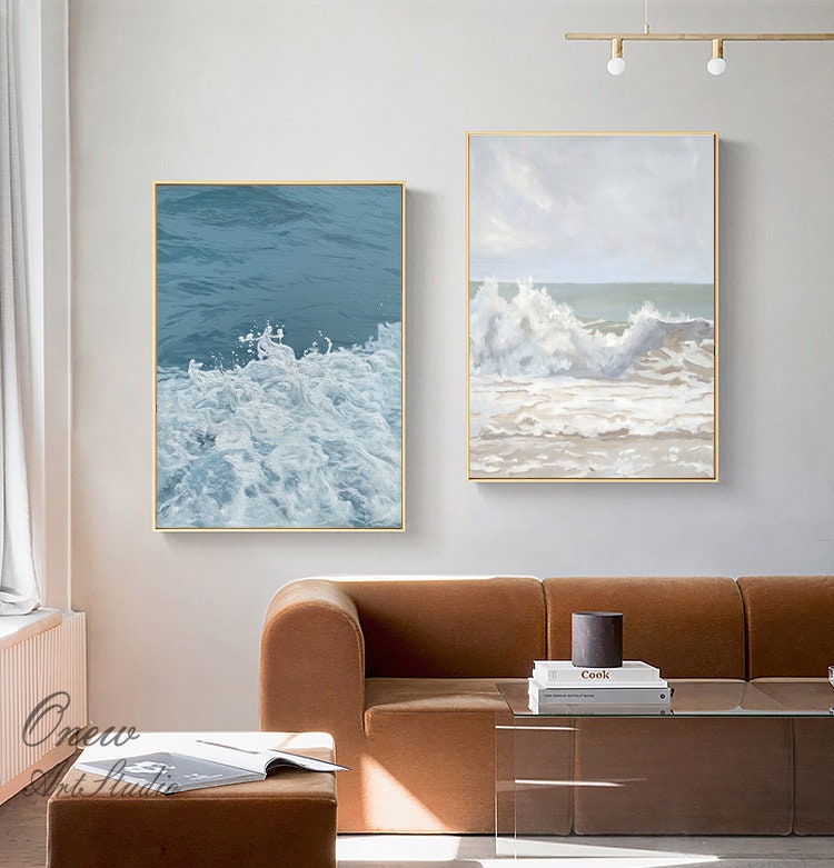 May include: Two abstract paintings of ocean waves. The painting on the left shows a close-up of white foamy waves against a blue background. The painting on the right shows a larger wave breaking on a sandy beach with a cloudy sky.