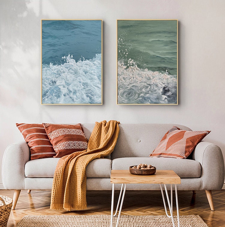 May include: Two framed abstract paintings of ocean waves with white foam. The paintings are hanging on a white wall above a light gray sofa with orange and white throw pillows and a yellow blanket.