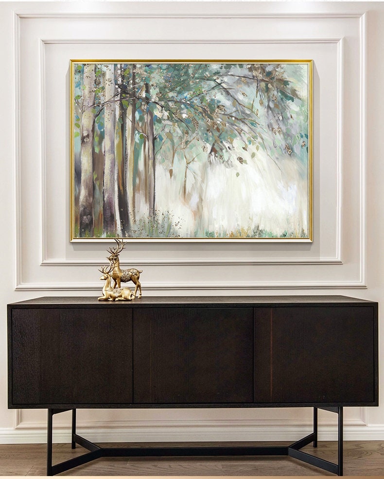 May include: A framed painting of a forest scene with trees and green foliage. The painting is hanging on a white wall above a dark brown wooden cabinet with three doors. A gold deer figurine is sitting on top of the cabinet.