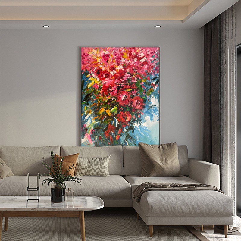 May include: A large abstract painting of a bouquet of pink, red, and yellow flowers with green leaves against a blue background. The painting is hanging on a wall in a living room with a gray sectional sofa and a coffee table.