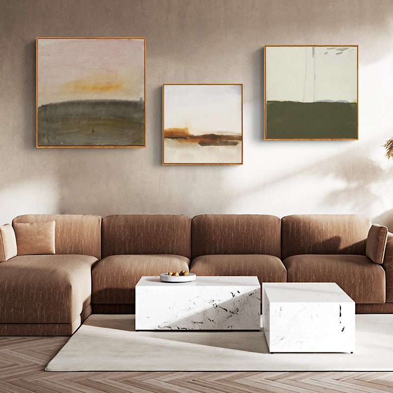 May include: Three abstract landscape paintings with muted colors in gold frames hanging on a beige wall above a brown sectional sofa and two white marble coffee tables.