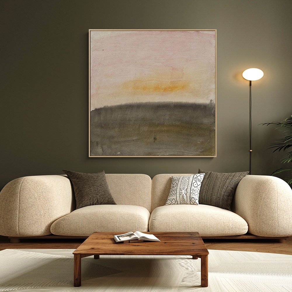 May include: A beige sofa with two pillows, a wooden coffee table with a book on it, and a framed abstract painting of a sunset over a field in shades of pink, yellow, and brown hanging on a green wall.