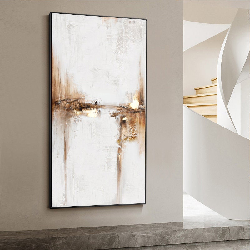 May include: Abstract painting with brown and white hues. The painting features a textured, layered style with subtle gold accents. The painting is framed in a black frame.