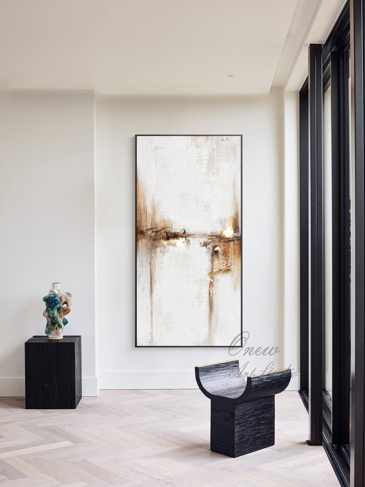 May include: Abstract painting in shades of brown, white, and gold, framed in black, hanging on a white wall in a modern interior. A black wooden stool with a curved top sits in the foreground.