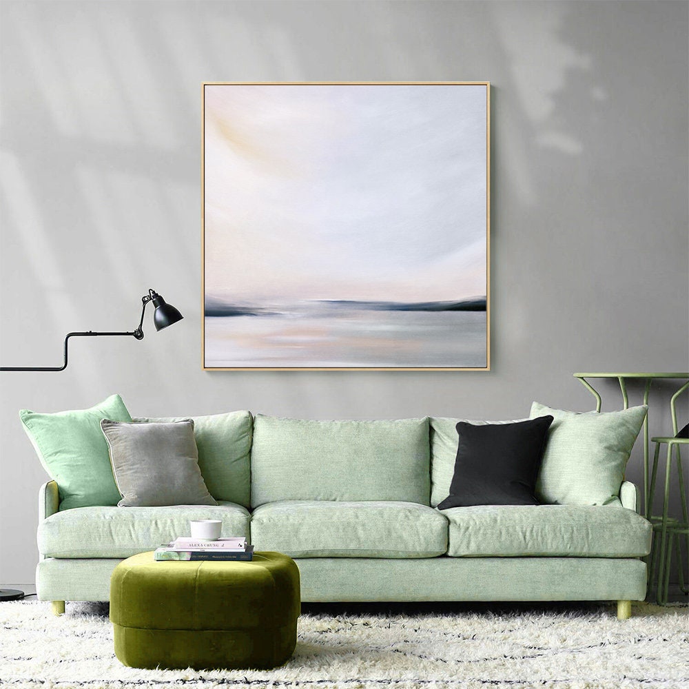 May include: A light green sofa with three pillows, a green ottoman, and a framed abstract painting of a seascape in muted tones of gray, white, and pink.