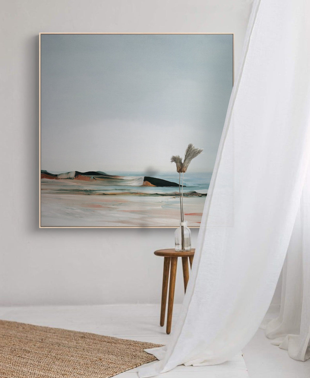 May include: A framed abstract landscape painting with a muted color palette of blues, greens, and browns. The painting depicts a beach scene with a distant shoreline and a cloudy sky. The painting is hanging on a white wall with a small wooden side table and a vase with dried flowers in front of it.
