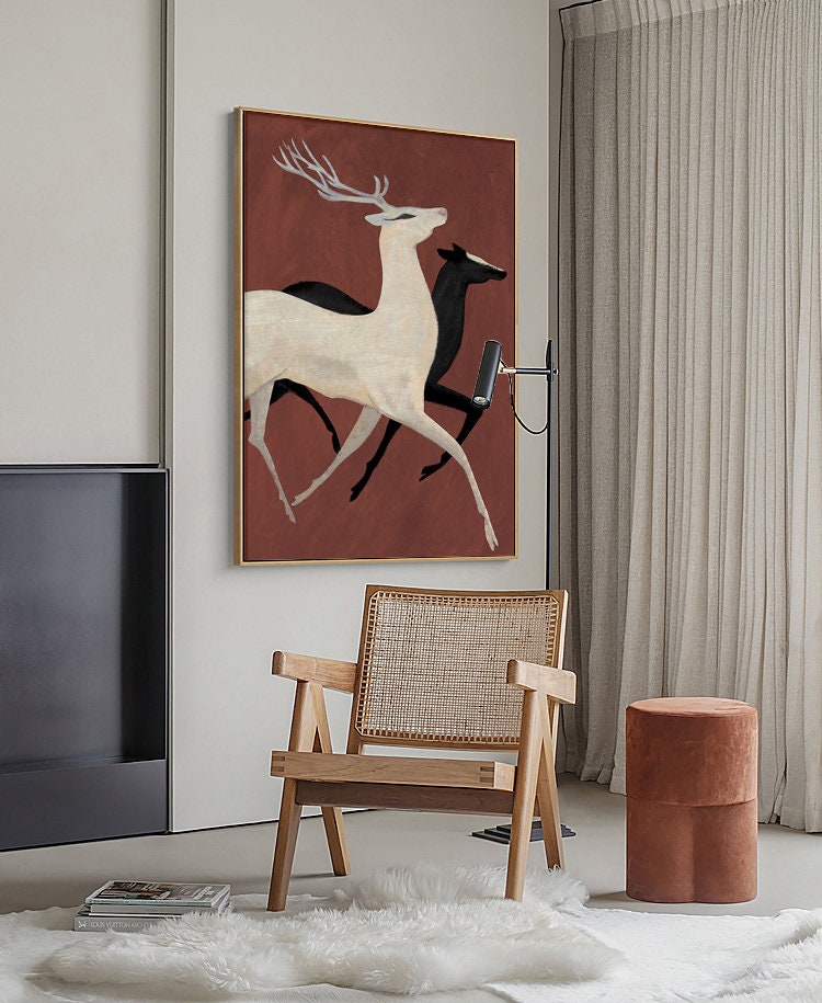 May include: A painting of two deer running, one white and one black, on a red background. The painting is framed in a gold frame and is hanging on a wall in a living room.