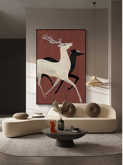 May include: A framed painting of two deer running on a red background. The deer are white and black, and the painting is hanging on a wall in a living room setting.