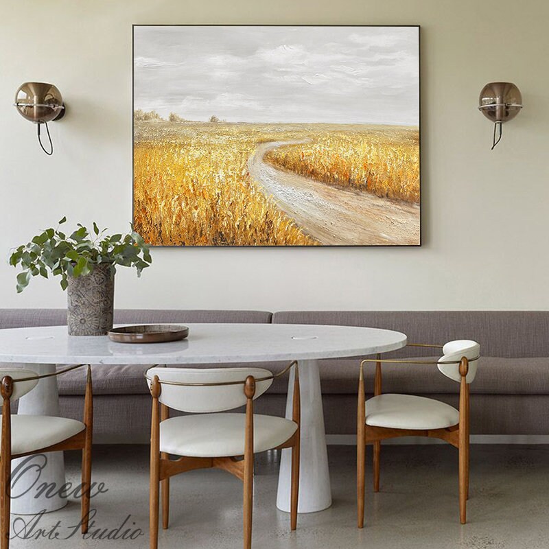 May include: A painting of a winding dirt road leading through a field of golden wheat. The sky is a soft gray with white clouds. The painting is framed and hanging on a wall above a white table and chairs.