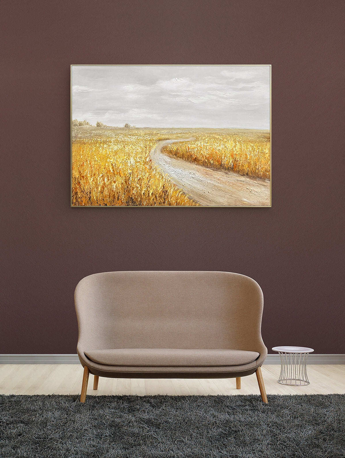 May include: A beige fabric sofa with a curved back and wooden legs sits in front of a brown wall with a framed landscape painting above it. The painting depicts a dirt road winding through a field of golden wheat.