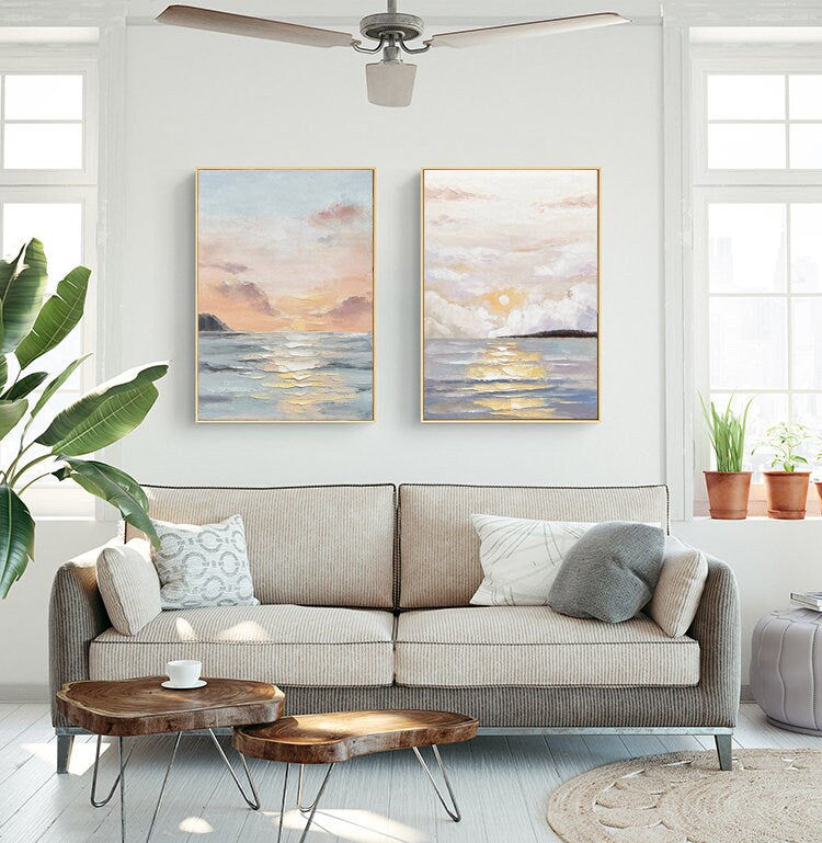 May include: Two framed paintings of seascapes with a sunset over the ocean. The paintings are in a living room setting with a couch, coffee table, and plants.