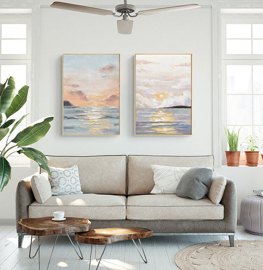 May include: Two framed paintings of seascapes with a sunset over the ocean. The paintings are in a living room setting with a couch, coffee table, and plants.