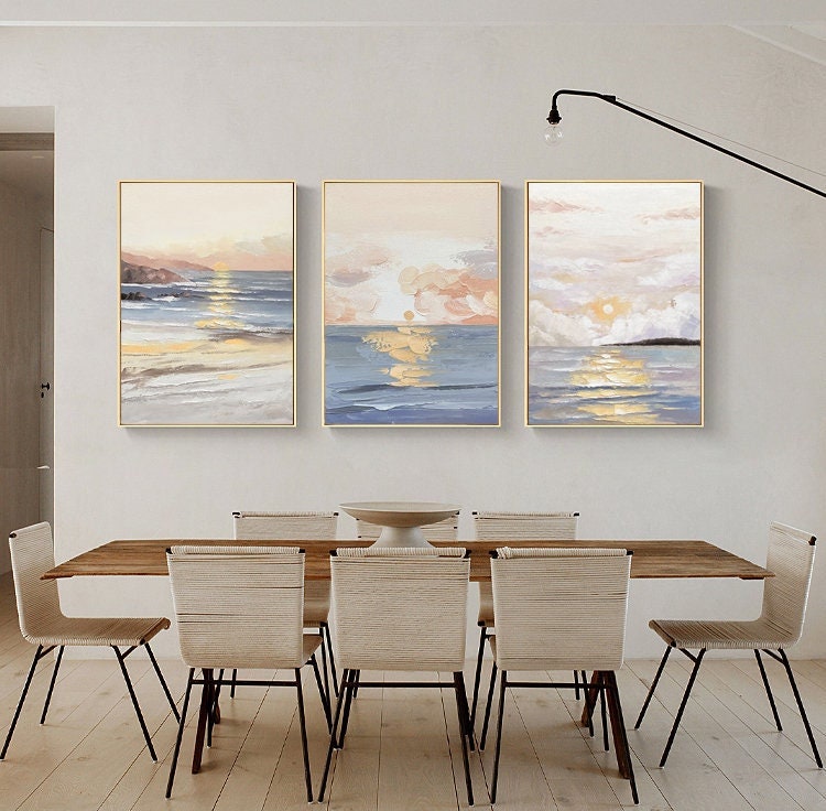 May include: Three abstract paintings of seascapes with a sunset over the ocean. The paintings are in shades of blue, pink, and yellow, and are framed in gold.