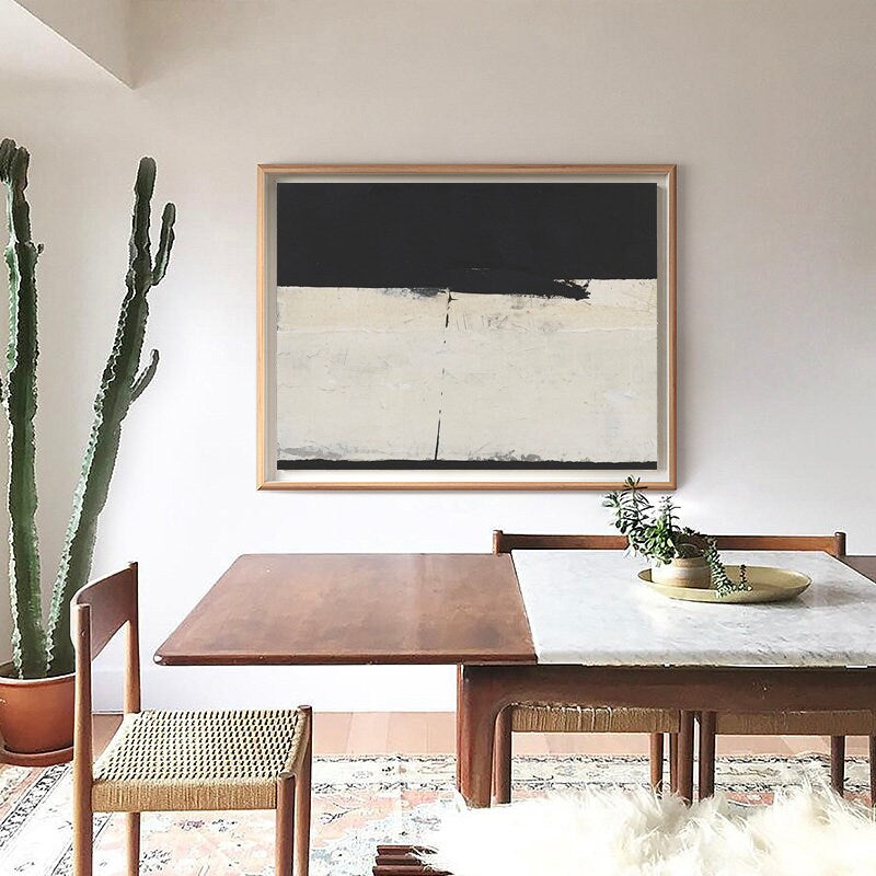 May include: A black and white abstract painting in a wooden frame hangs on a white wall above a wooden dining table with chairs. A cactus plant is in a terracotta pot on the left side of the image.