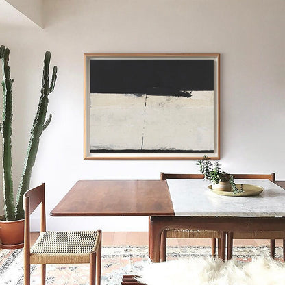 May include: A black and white abstract painting in a wooden frame hangs on a white wall above a wooden dining table with chairs. A cactus plant is in a terracotta pot on the left side of the image.