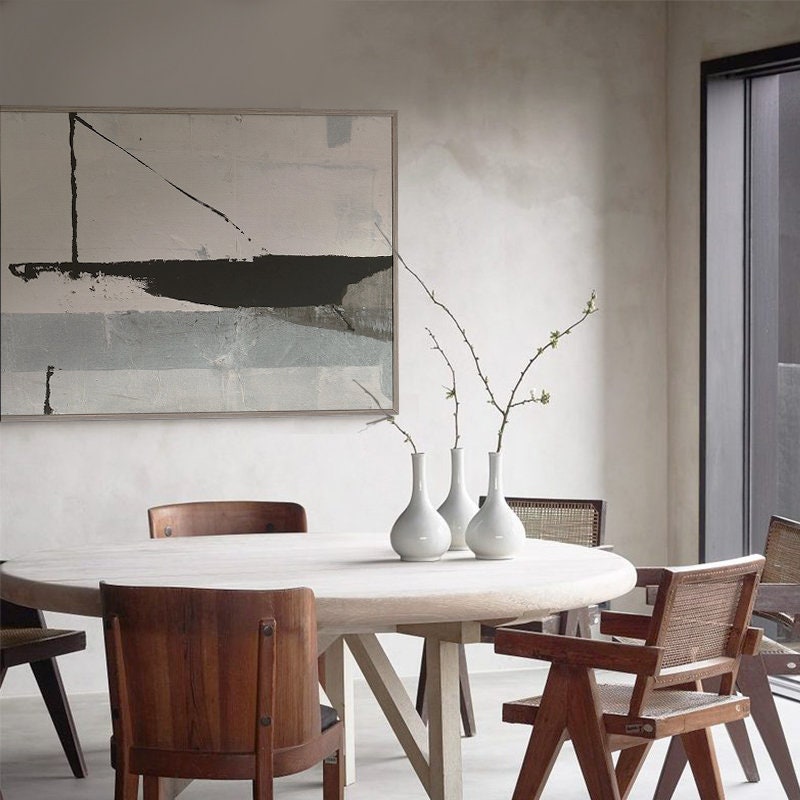 May include: A round wooden dining table with a white top and four chairs. The chairs are made of wood with a brown finish. There is a white abstract painting on the wall behind the table. A vase with three branches of white flowers sits on the table.