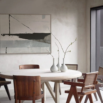 May include: A round wooden dining table with a white top and four chairs. The chairs are made of wood with a brown finish. There is a white abstract painting on the wall behind the table. A vase with three branches of white flowers sits on the table.