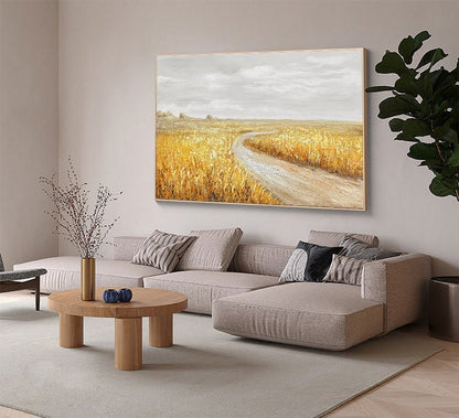 May include: A painting of a winding dirt road leading through a field of golden wheat. The sky is a soft gray with white clouds. The painting is framed in a light wood frame.