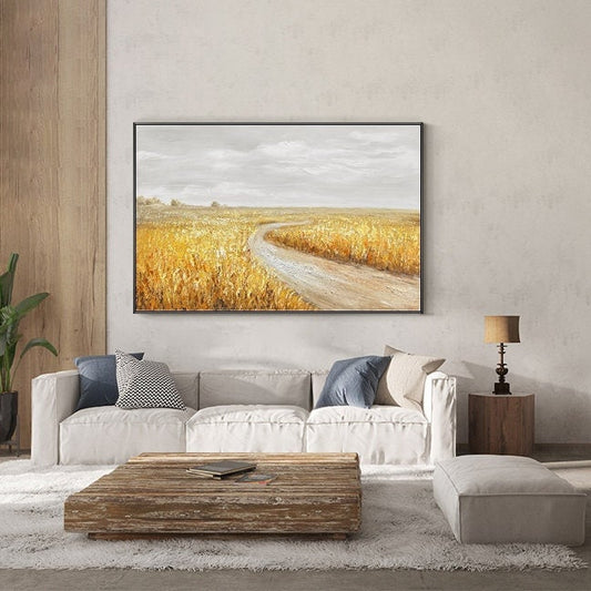 May include: A framed canvas painting of a dirt road winding through a field of golden wheat. The sky is a light gray with a few clouds. The painting is hanging on a wall above a white couch.