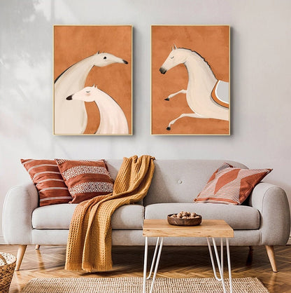 May include: Two abstract paintings of white horses on a burnt orange background. The paintings are framed in gold and hung on a white wall.