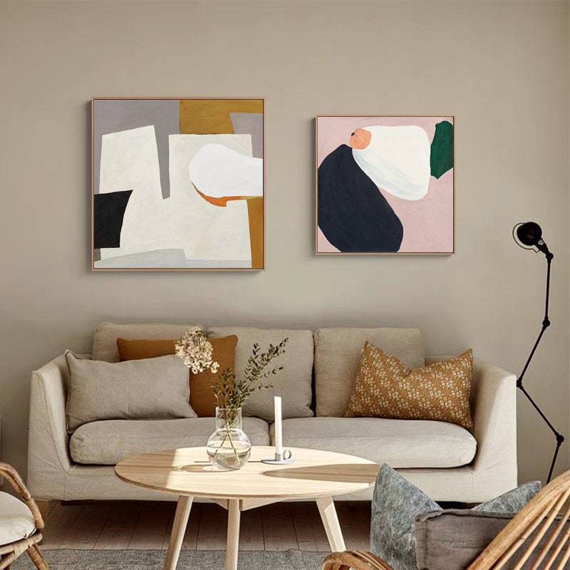 May include: Two abstract paintings in square frames, one with white, gray, black, and orange shapes, the other with white, pink, green, and blue shapes, hanging on a beige wall above a light beige sofa with two pillows.