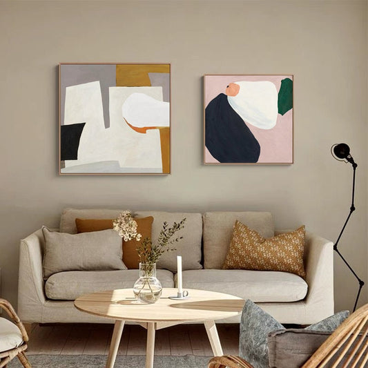 May include: Two abstract paintings in square frames, one with white, gray, black, and orange shapes, the other with white, pink, green, and blue shapes, hanging on a beige wall above a light beige sofa with two pillows.