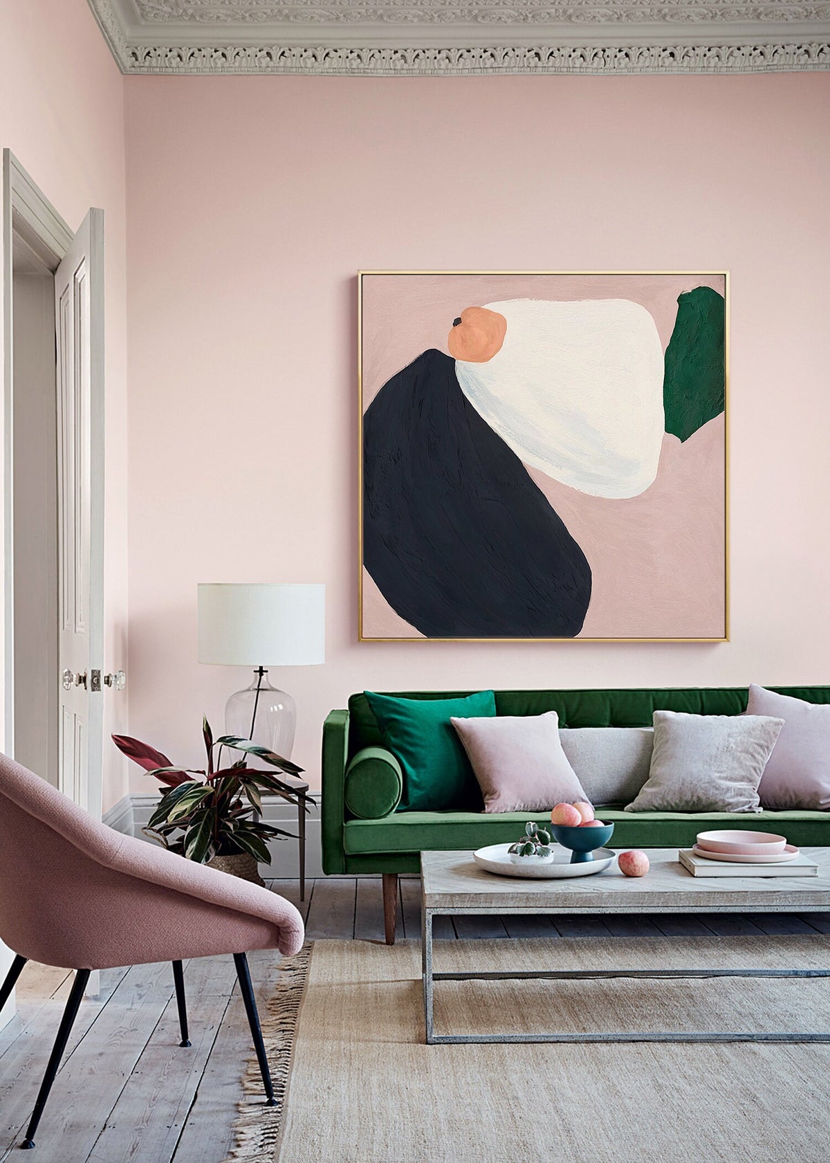 May include: A living room with a green velvet sofa, pink armchair, and a coffee table with a bowl of fruit. The walls are painted a pale pink and there is a large abstract painting on the wall.