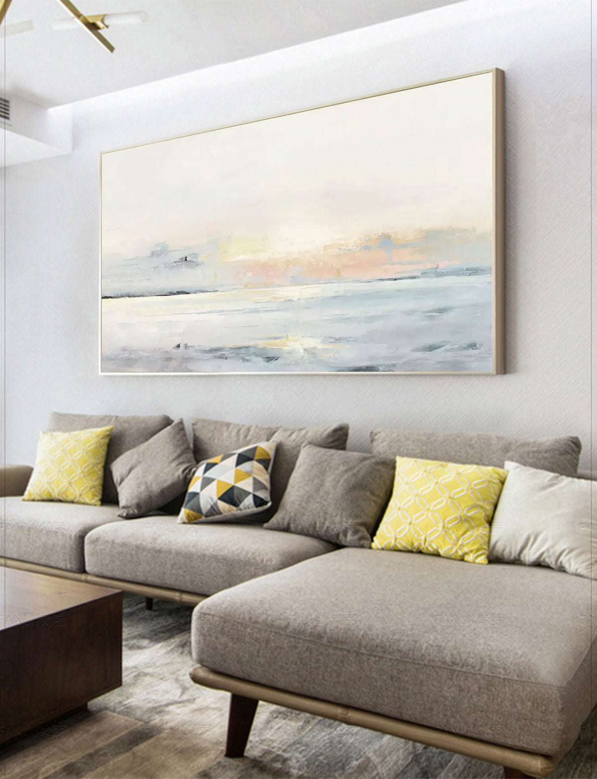 May include: Large abstract landscape painting in a gold frame. The artwork features a soft color palette of pale blues, pinks, and yellows, evoking a serene coastal scene. The painting hangs above a gray sectional sofa with decorative pillows.