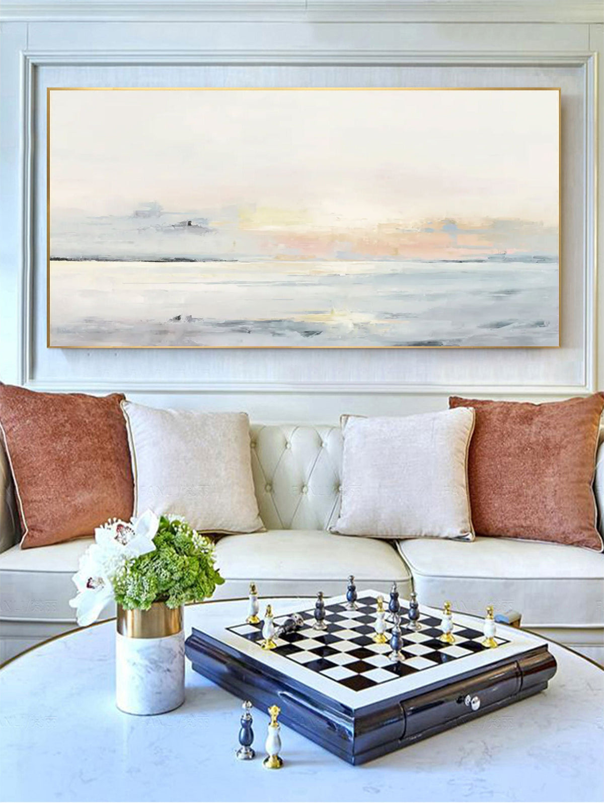May include: An abstract landscape painting in a gold frame hangs above a white sofa with decorative pillows. A chess set with black and white pieces sits on a table, next to a vase of flowers.