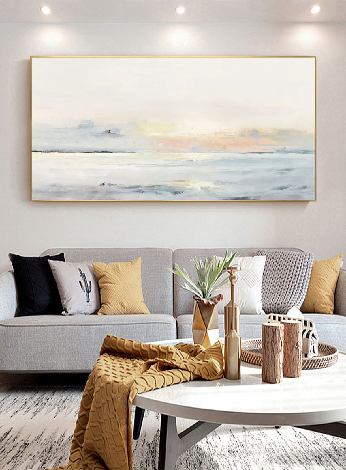 May include: A large rectangular abstract painting with a gold frame depicts a serene seascape in muted tones. The artwork hangs above a gray sofa adorned with pillows in black, yellow, and white. A coffee table displays decorative items.