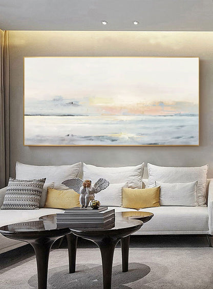 May include: An abstract landscape painting in a gold frame hangs above a white sofa. The artwork depicts a serene seascape with soft blues, whites, and a hint of peach. A small angel statue sits on a stack of books on a black table.