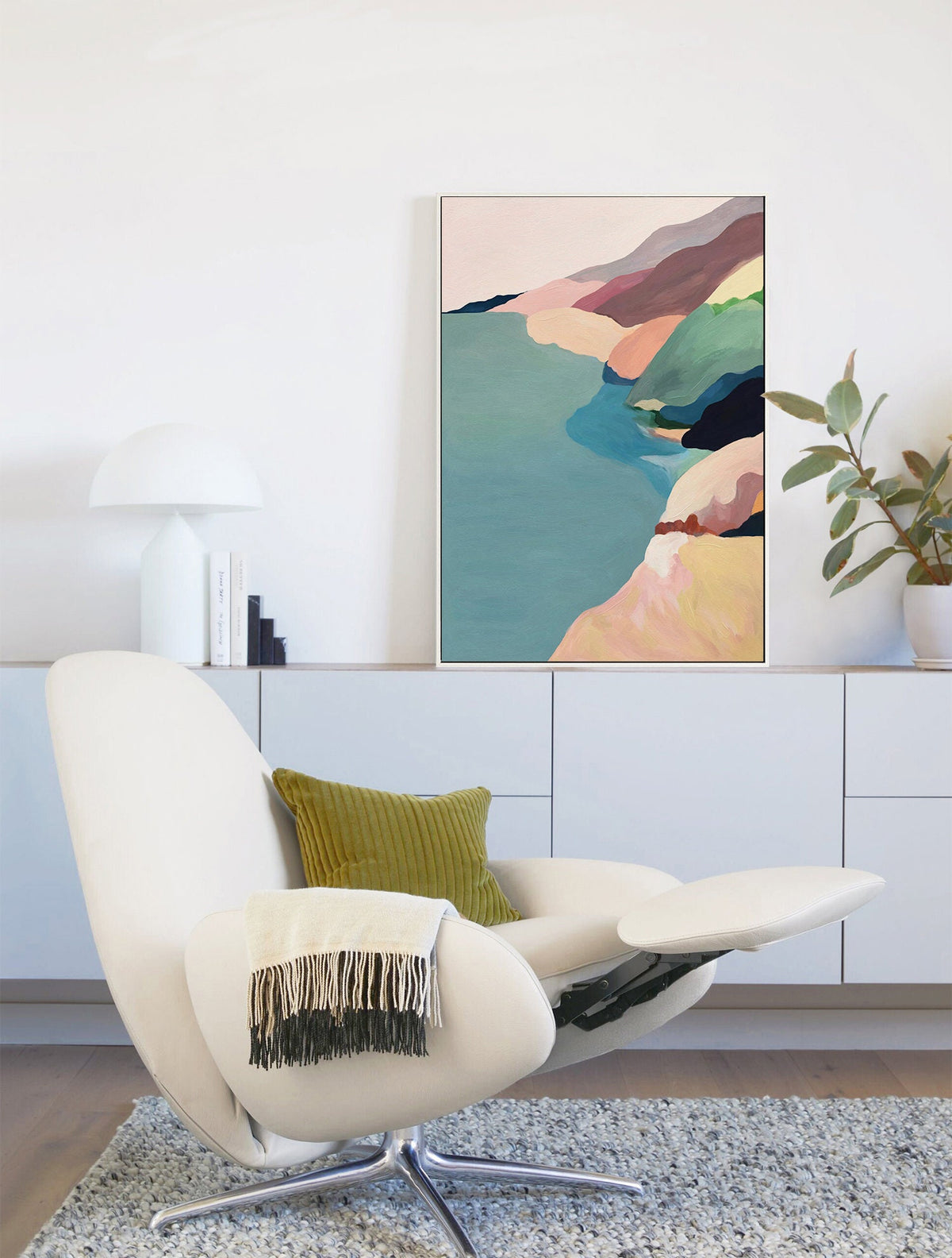 May include: A white armchair with a green pillow and a white blanket with fringe. The armchair is in front of a white wall with a framed abstract painting of a coastal landscape in shades of pink, blue, green, and brown.