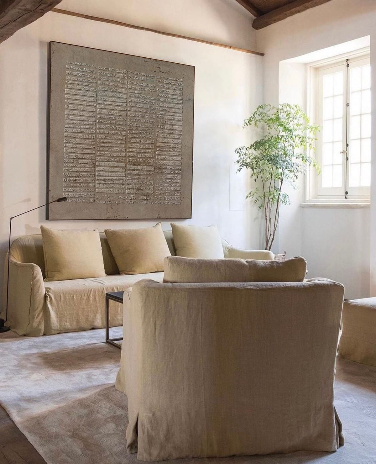 May include: A large square wall art piece featuring a textured, grid-like pattern in shades of gray and beige, displayed above a beige slipcovered sofa with three matching throw pillows and a matching armchair in a bright, open living space.